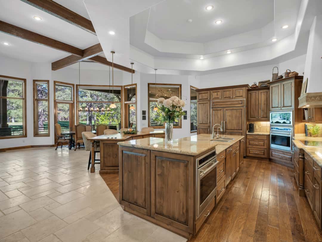 9 of 52. Open-concept great room with soaring ceilings, exposed wood beams, fireplace feature wall, and expansive windows overlooking scenic pine forest surroundings.