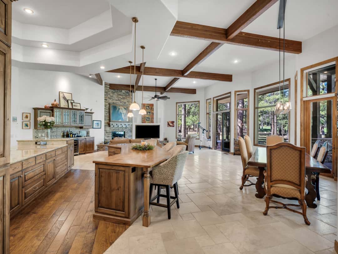 12 of 52. Open-concept kitchen and dining area with exposed beams, large windows, custom wood cabinetry, and seamless connection to the surrounding pine forest views.