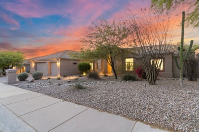 Inviting front exterior showcasing mature desert landscaping, modern architecture, and a spacious three-car garage. The professionally designed landscape enhances the home's impressive curb appeal and low-maintenance lifestyle.