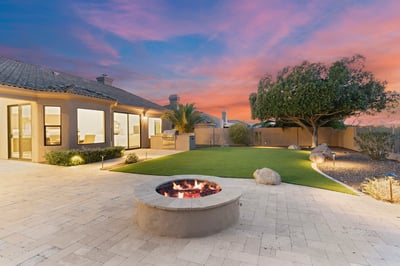 Twilight backyard featuring a glowing fire pit, outdoor kitchen, professionally landscaped yard, and spacious travertine patio. Experience luxury outdoor living in Scottsdale.