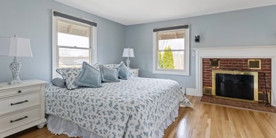 This inviting bedroom features large windows, cozy furnishings, and a warm brick fireplace, perfect for relaxation.