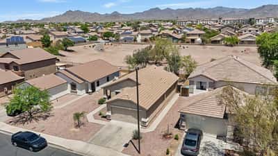 Bird's-eye view captures the home's front elevation, surrounding neighborhood, and nearby open space in Buckeye. This aerial photo emphasizes the property's desirable setting, low-maintenance landscaping, mountain backdrop, and easy access to parks and community amenities while showcasing the home's inviting curb appeal.