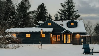 Cozy home surrounded by snow-covered trees, featuring warm interior lighting and inviting outdoor space.