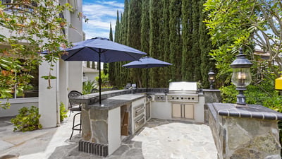 Discover this inviting outdoor kitchen space withViking BBQ, Cooktop, Warming Drawers, outdoor lighting and shady umbrellas, perfect for entertaining, surrounded by lush greenery and ample seating.
