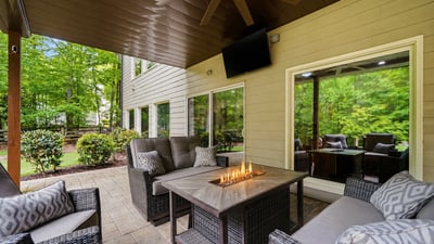 A covered outdoor living area designed for year-round enjoyment complete with a fireside table, TV, ceiling fan, and wooded views that make the space feel private, comfortable, and easy to enjoy.