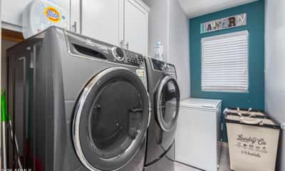 This dedicated laundry room features upper cabinetry for storage, modern laundry appliances, and a window providing natural light. A practical and organized utility space enhances everyday convenience.