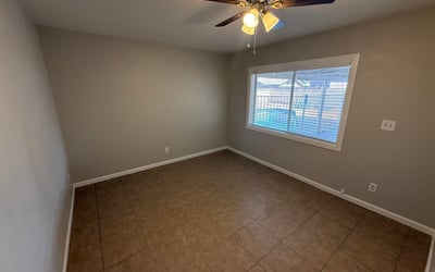 Guest bedroom with tile flooring, neutral colors, and ceiling fan for comfort. Large window allows natural light while maintaining a calm and inviting atmosphere.