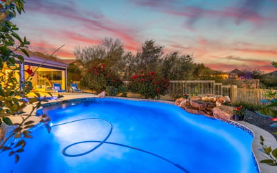 Scottsdale backyard with glowing pool, waterfall feature, and vibrant sunset backdrop, perfect for relaxing evenings outdoors.