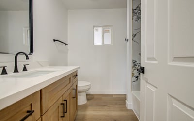 This renovated bathroom features a quartz vanity, custom cabinetry, matte black fixtures, and a striking marble-look tub and shower surround for everyday luxury.