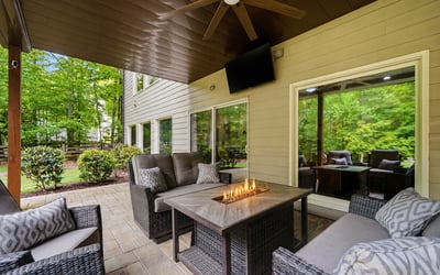 A covered outdoor living area designed for year-round enjoyment complete with a fireside table, TV, ceiling fan, and wooded views that make the space feel private, comfortable, and easy to enjoy.