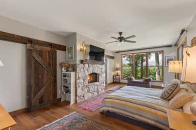 Cozy bedroom featuring a stone fireplace, large windows with scenic views, and rustic wood accents.
