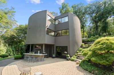 This distinctive modern home features large windows and a unique curved design, surrounded by lush landscaping.