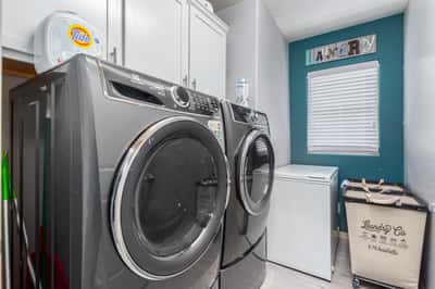 This dedicated laundry room features upper cabinetry for storage, modern laundry appliances, and a window providing natural light. A practical and organized utility space enhances everyday convenience.