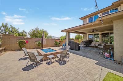 Alternate backyard view showcases the inviting covered patio, fire pit seating area, paver hardscape, and colorful landscaping designed for comfortable outdoor living.
