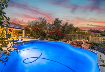 Scottsdale backyard with glowing pool, waterfall feature, and vibrant sunset backdrop, perfect for relaxing evenings outdoors.