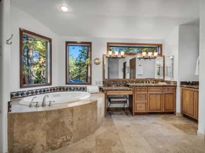 Luxurious primary bathroom with Jacuzzi soaking tub, dual sink vanity, natural stone finishes, and oversized windows with forest views. This spa-inspired space offers tranquility and refined mountain elegance.