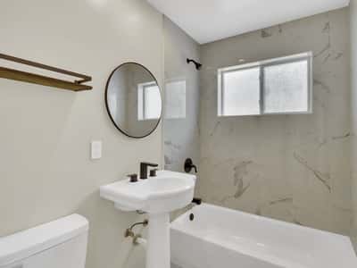 Fully renovated hall bathroom featuring marble-look tile walls, modern black fixtures, a pedestal sink, and abundant natural light. Stylish finishes add lasting appeal to this Phoenix rental.