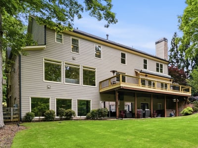 Spacious home featuring expansive windows and a large deck, nestled among lush greenery and mature trees.