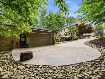 Spacious driveway with natural landscaping offers a welcoming approach to this unique property surrounded by trees.