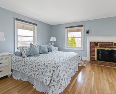 This inviting bedroom features large windows, cozy furnishings, and a warm brick fireplace, perfect for relaxation.