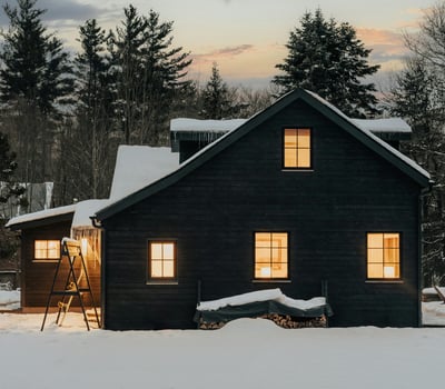 Cozy black cabin with warm lights, surrounded by snow and towering trees, perfect for winter getaways.