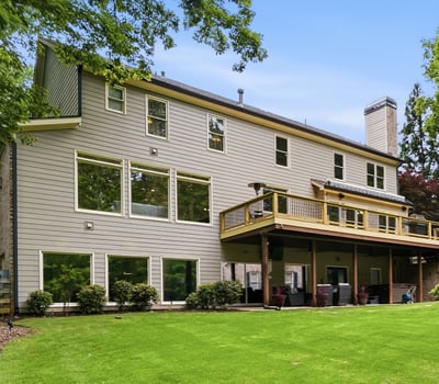 Spacious home featuring expansive windows and a large deck, nestled among lush greenery and mature trees.