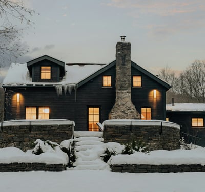 This cozy black-clad home features a stone chimney and inviting outdoor spaces, nestled in a winter landscape.