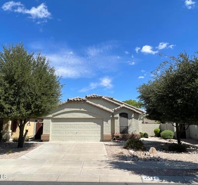 Welcoming front elevation framed by mature trees and desert landscaping for low-maintenance living. The spacious driveway and attached garage provide convenience while complementing the home's appealing design.
