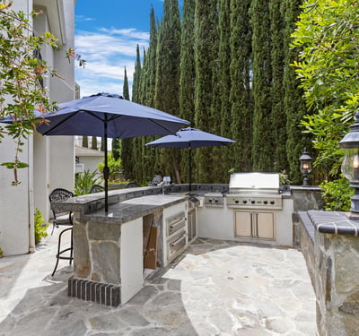 Discover this inviting outdoor kitchen space withViking BBQ, Cooktop, Warming Drawers, outdoor lighting and shady umbrellas, perfect for entertaining, surrounded by lush greenery and ample seating.