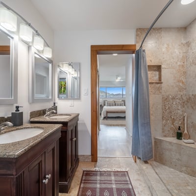 Spacious dual-sink bathroom features a stone-tiled shower and a view of the adjoining bedroom.