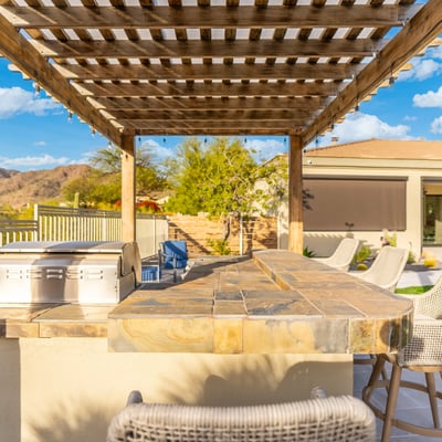 Scottsdale backyard kitchen with stone counters, grill, and seating under pergola, ideal for gatherings and outdoor dining.