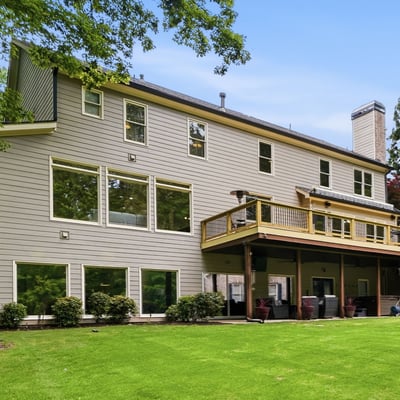 Spacious home featuring expansive windows and a large deck, nestled among lush greenery and mature trees.