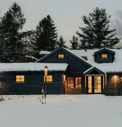 Cozy home surrounded by snow-covered trees, featuring warm interior lighting and inviting outdoor space.