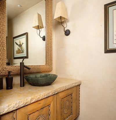Thoughtfully designed with a carved wood cabinet, stone-style countertop, and a green glass vessel sink, this half bath adds unique character to the home. Rope detailing around the mirror and warm wall tones give it a rustic yet refined look.