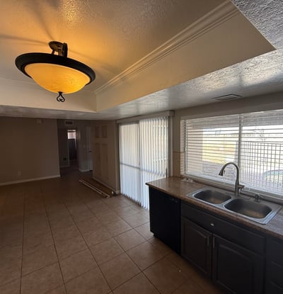 Kitchen area with double sink, tile counters, and rich cabinetry. Natural light from the wide window brightens the room while offering a glimpse of the pool area outside.