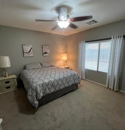 Large primary bedroom with plush carpet, ceiling fan, and oversized windows that bring in natural light, creating a peaceful and inviting space to unwind.