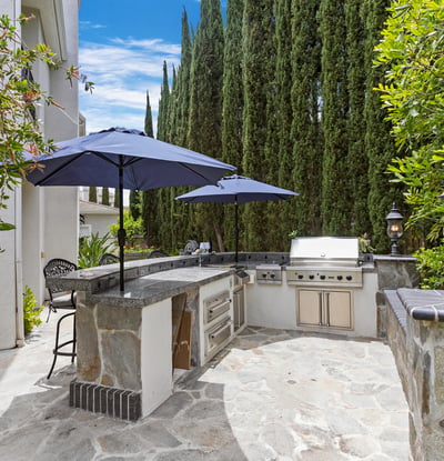 Discover this inviting outdoor kitchen space withViking BBQ, Cooktop, Warming Drawers, outdoor lighting and shady umbrellas, perfect for entertaining, surrounded by lush greenery and ample seating.