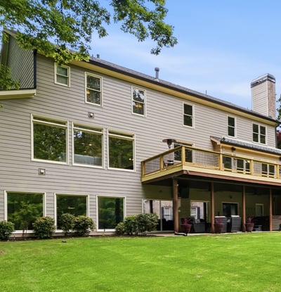 Spacious home featuring expansive windows and a large deck, nestled among lush greenery and mature trees.