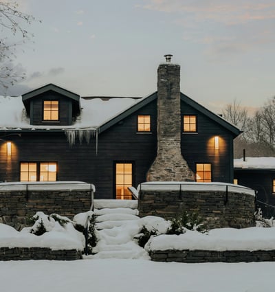 This cozy black-clad home features a stone chimney and inviting outdoor spaces, nestled in a winter landscape.