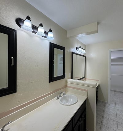 Functional primary bathroom featuring dual sinks, updated lighting, ample storage, and convenient access to a spacious walk-in closet. The thoughtful layout supports both comfort and everyday efficiency.