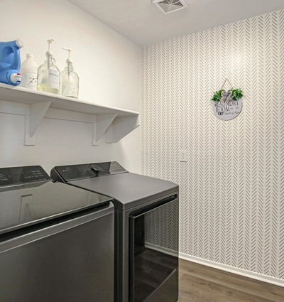 Well-appointed laundry room featuring stylish wallpaper, modern flooring, upper shelving, and dedicated space for laundry appliances. A practical upgrade for everyday living in this Waddell home.