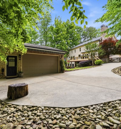 Spacious driveway with natural landscaping offers a welcoming approach to this unique property surrounded by trees.