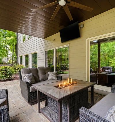 A covered outdoor living area designed for year-round enjoyment complete with a fireside table, TV, ceiling fan, and wooded views that make the space feel private, comfortable, and easy to enjoy.