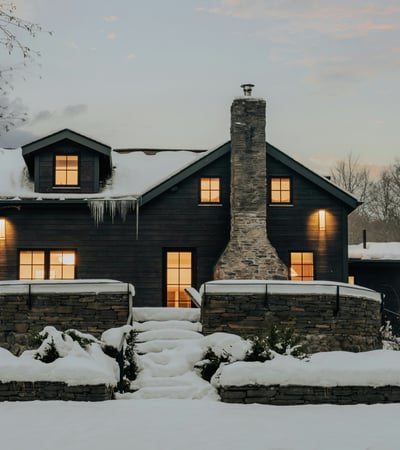 This cozy black-clad home features a stone chimney and inviting outdoor spaces, nestled in a winter landscape.