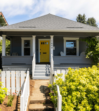 This inviting home features a bright yellow door, spacious porch, and vibrant landscaping in a friendly neighborhood.
