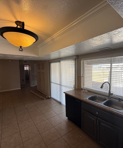 Kitchen area with double sink, tile counters, and rich cabinetry. Natural light from the wide window brightens the room while offering a glimpse of the pool area outside.