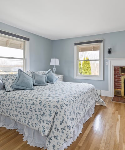 This inviting bedroom features large windows, cozy furnishings, and a warm brick fireplace, perfect for relaxation.