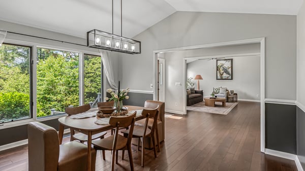 Bright, open dining area seamlessly connects to a spacious living room, framed by large windows showcasing lush views.