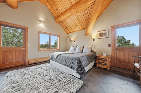 The bright bedroom features vaulted wood ceilings, large windows, and access to two outdoor balconies.