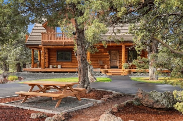 Beautifully crafted log cabin featuring a spacious porch, surrounded by lush greenery and outdoor seating.